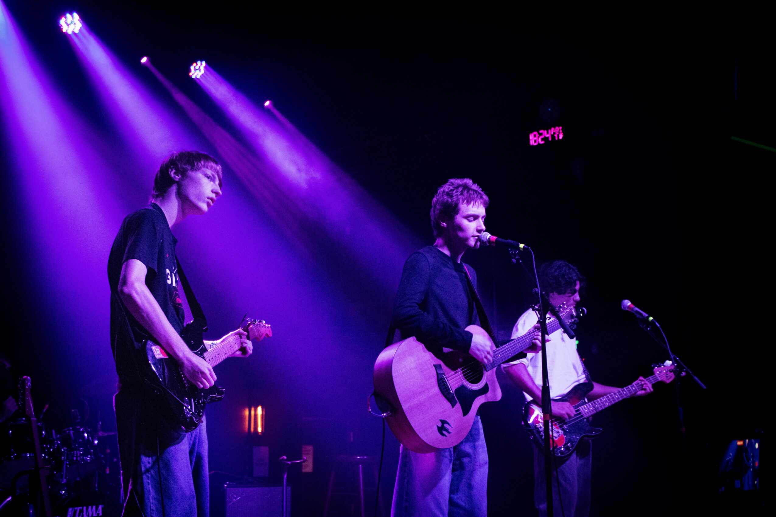 Three musicians perform on stage with purple lighting, one playing guitar and singing into a microphone.