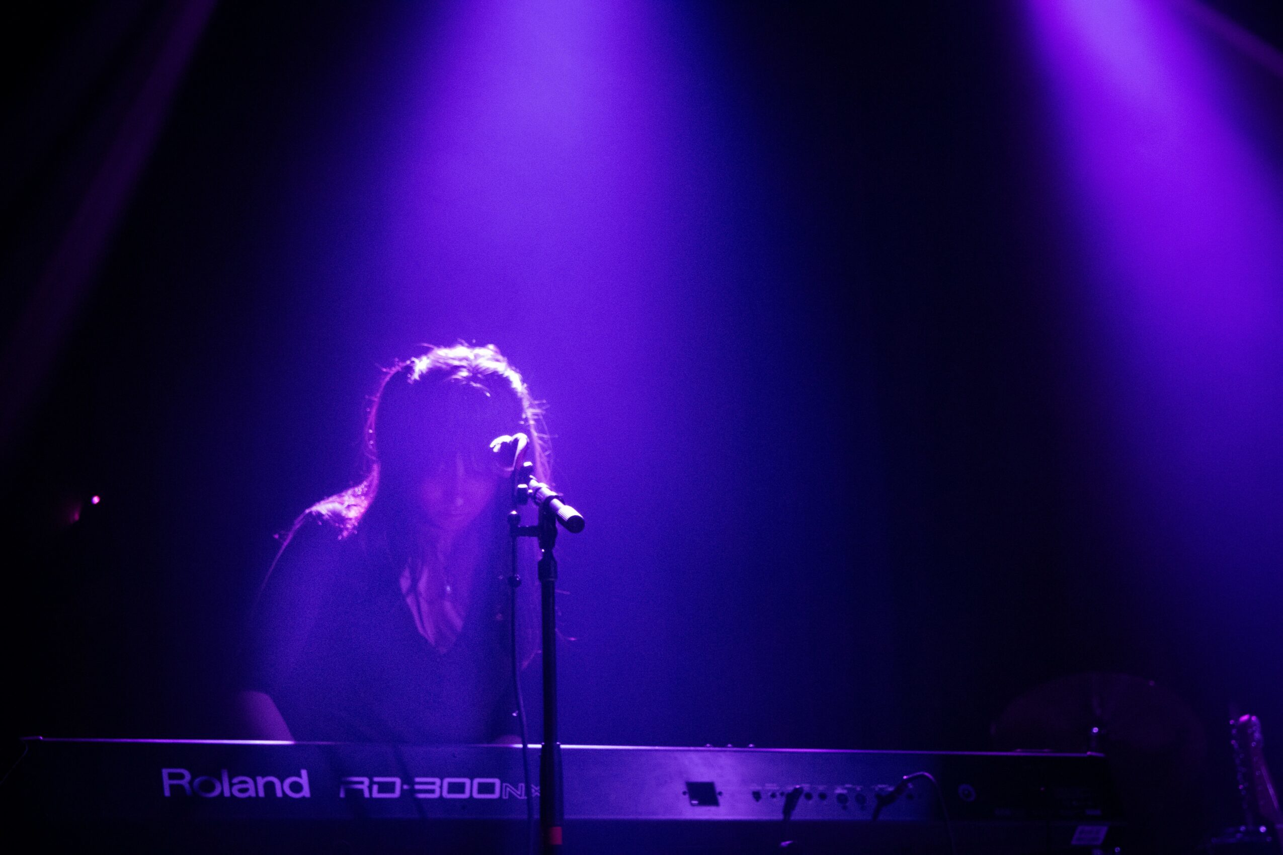 Person playing keyboard on stage with purple lighting, singing into microphone, dark background