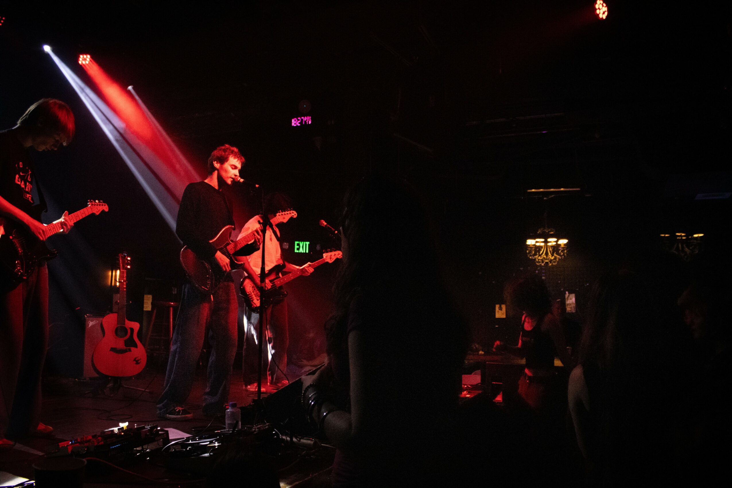 Musicians perform on stage with guitars and microphones, stage lighting creates a dark atmosphere, audience visible in shadows.