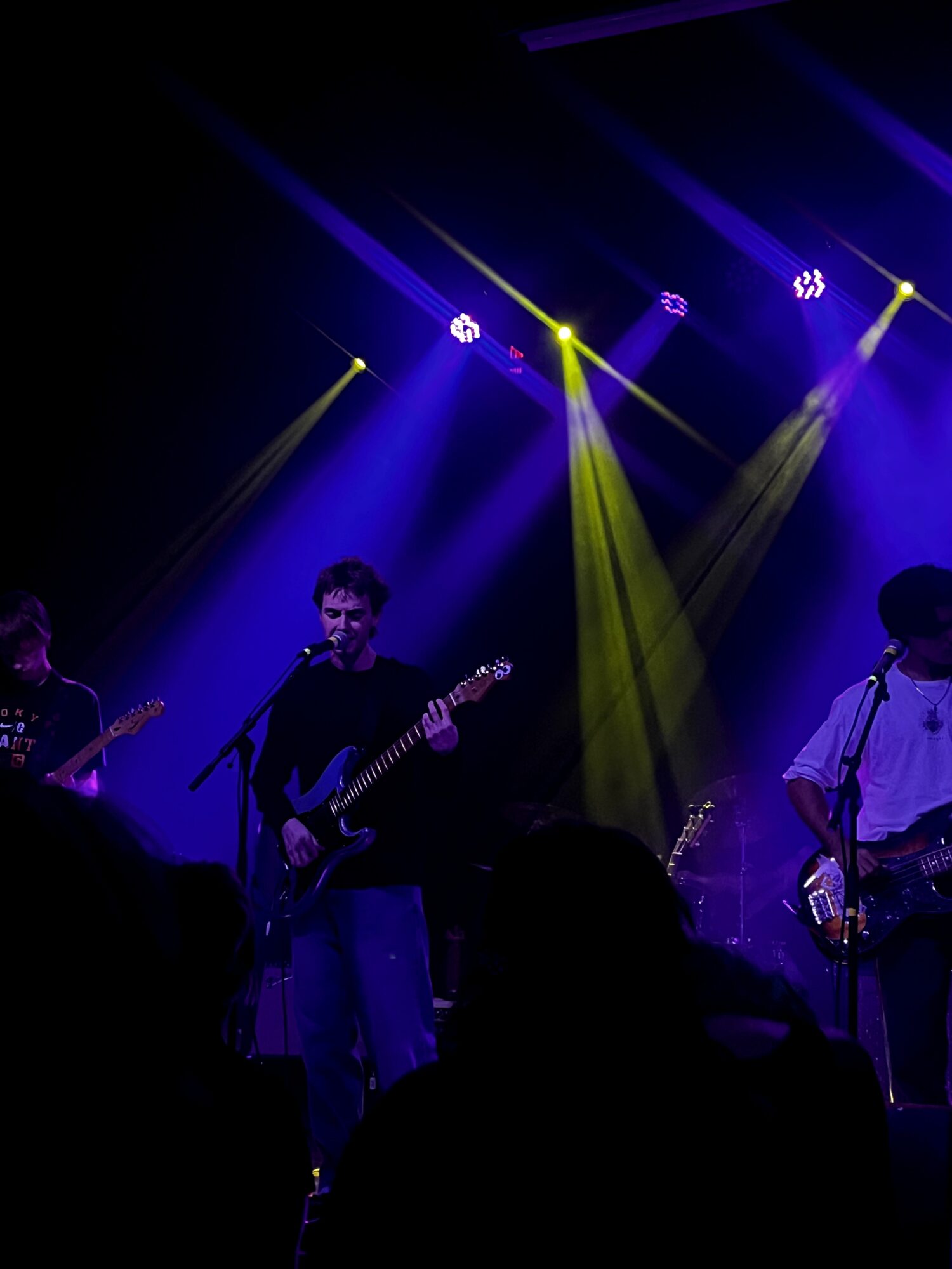 Band performs on stage with colorful lighting, three musicians playing guitars, audience silhouettes visible in foreground.