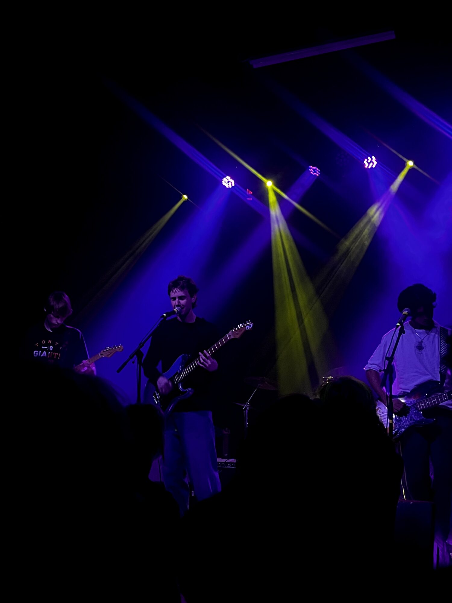 Band performing on stage with colorful lighting, three musicians playing guitars, audience silhouettes visible.