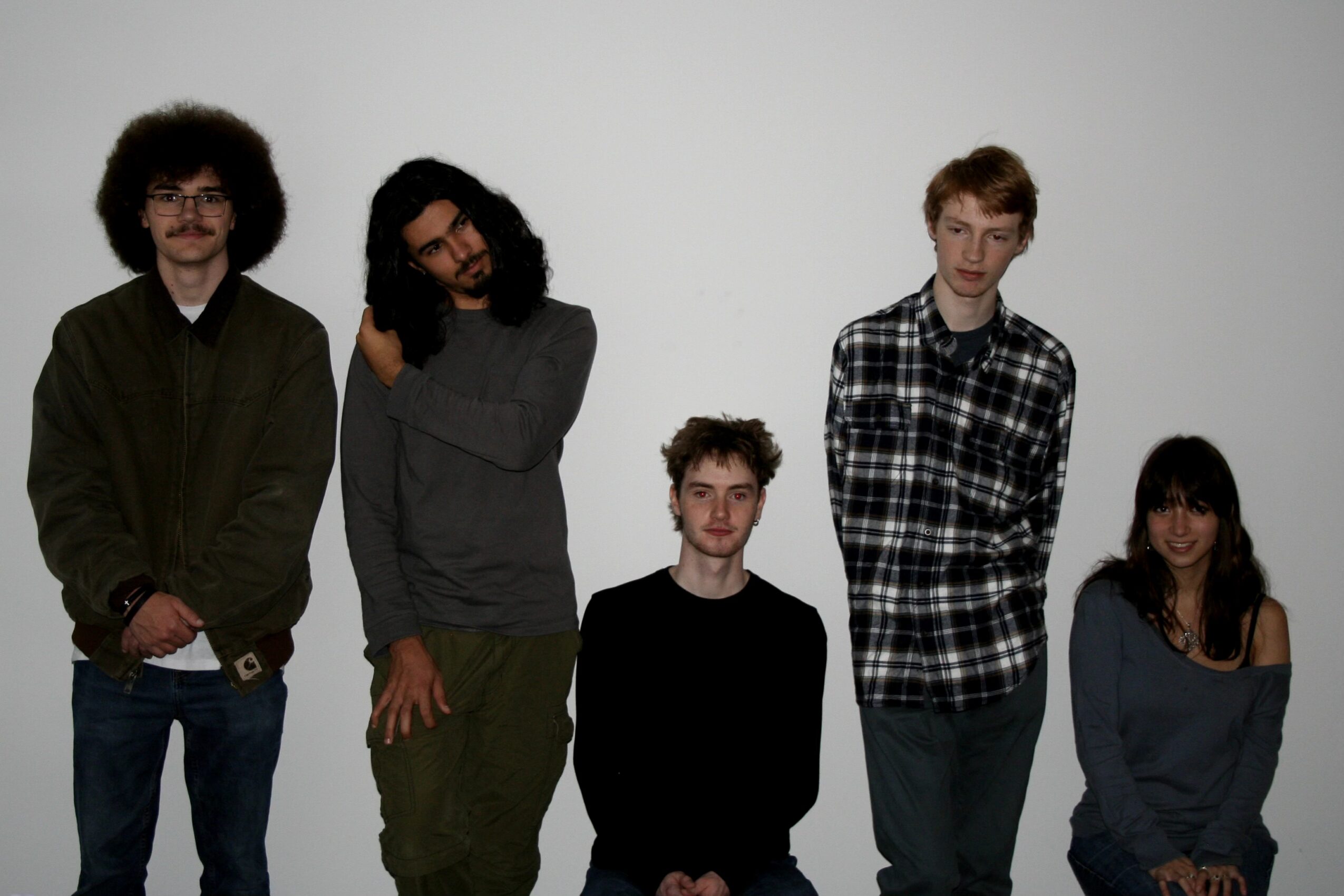 Five people standing in a row against a plain background, facing forward, with varied clothing styles and hair types.