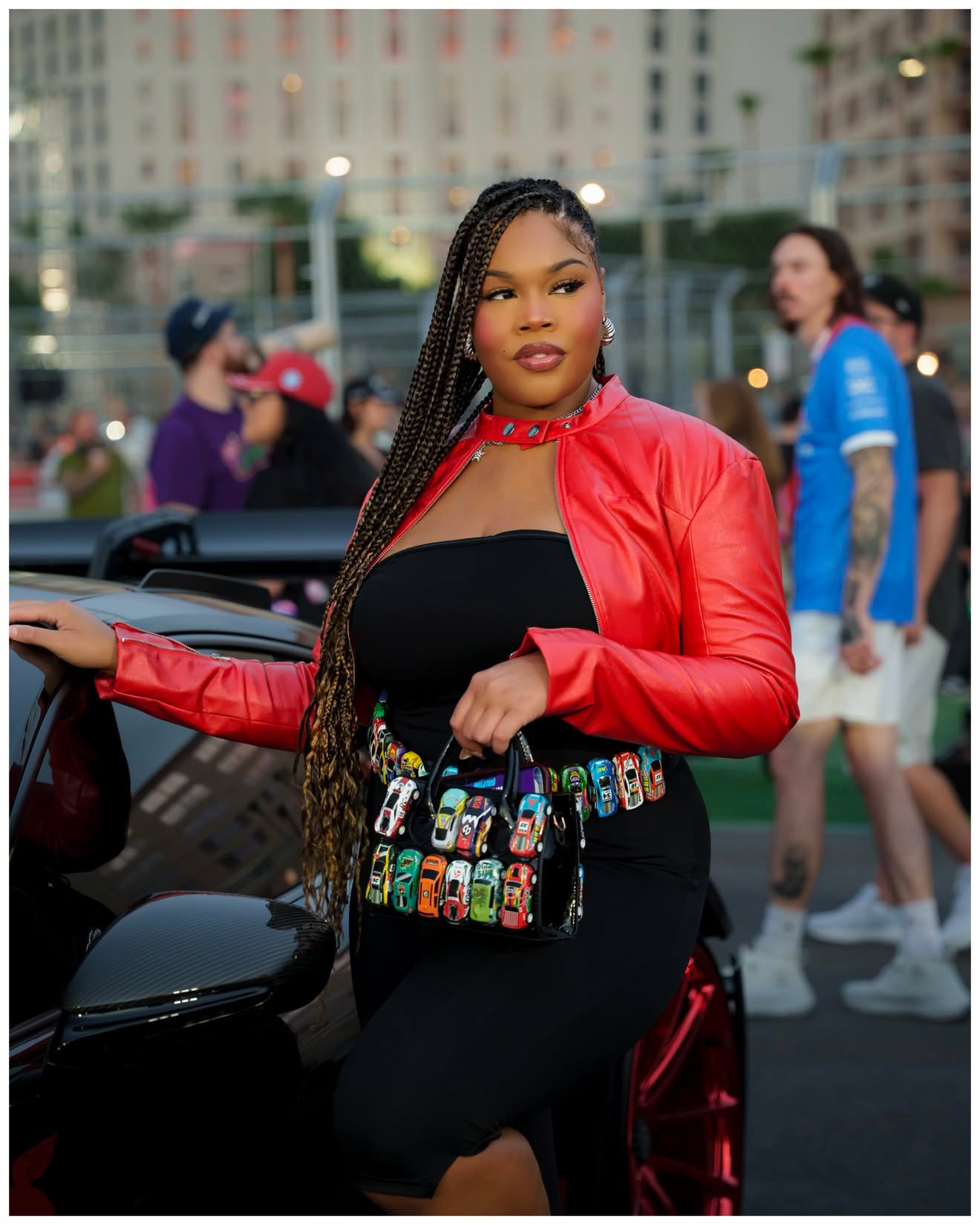 Woman with braided hair in a red jacket and black dress, holding a colorful bag, standing outdoors with people in the background.