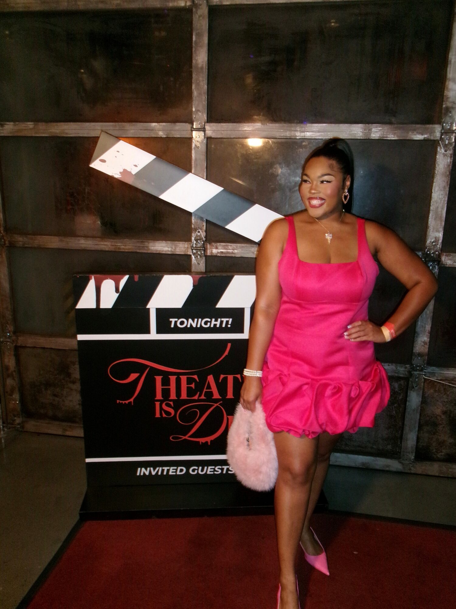Young woman in pink dress standing next to a movie-themed sign, smiling, with a dark background.