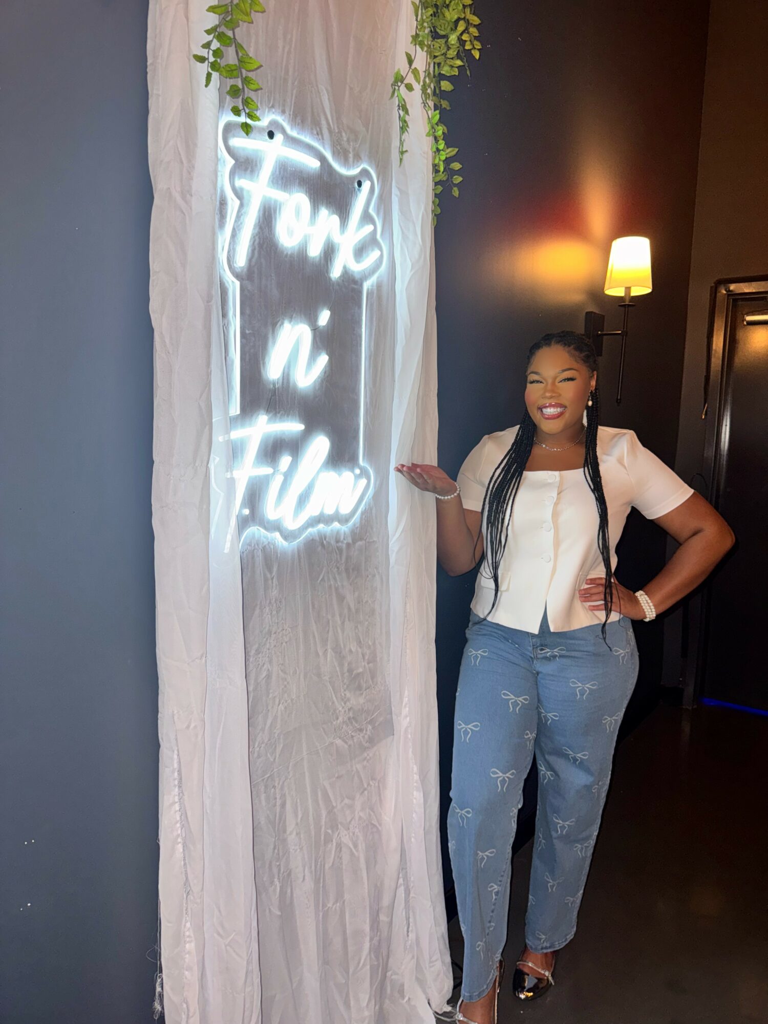 Woman with long braids smiling, standing next to a neon sign that reads 'Funk 'n' Flava' in a dimly lit room.