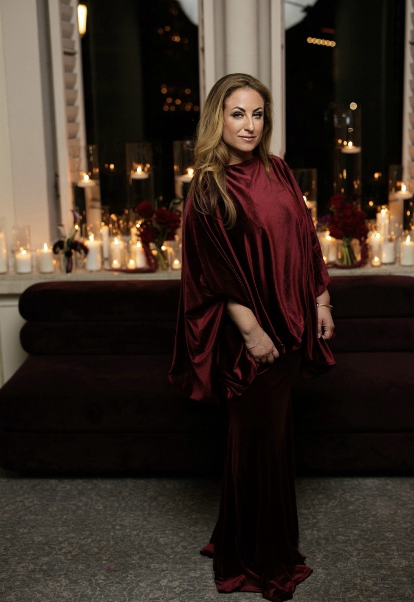 Woman in a red velvet dress standing indoors at night with lit candles and city lights visible through windows.