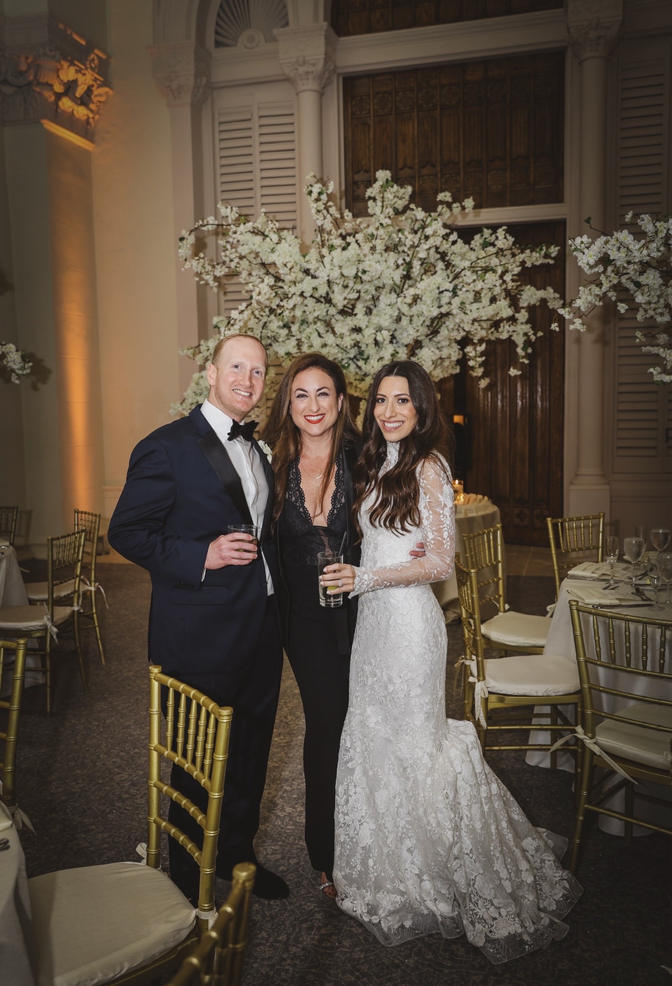 Three people in formal attire standing in a decorated event space with floral arrangements and tables.