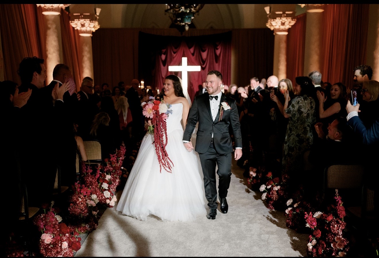 Bride and groom walking down aisle with guests and a cross in background, floral decorations on sides.
