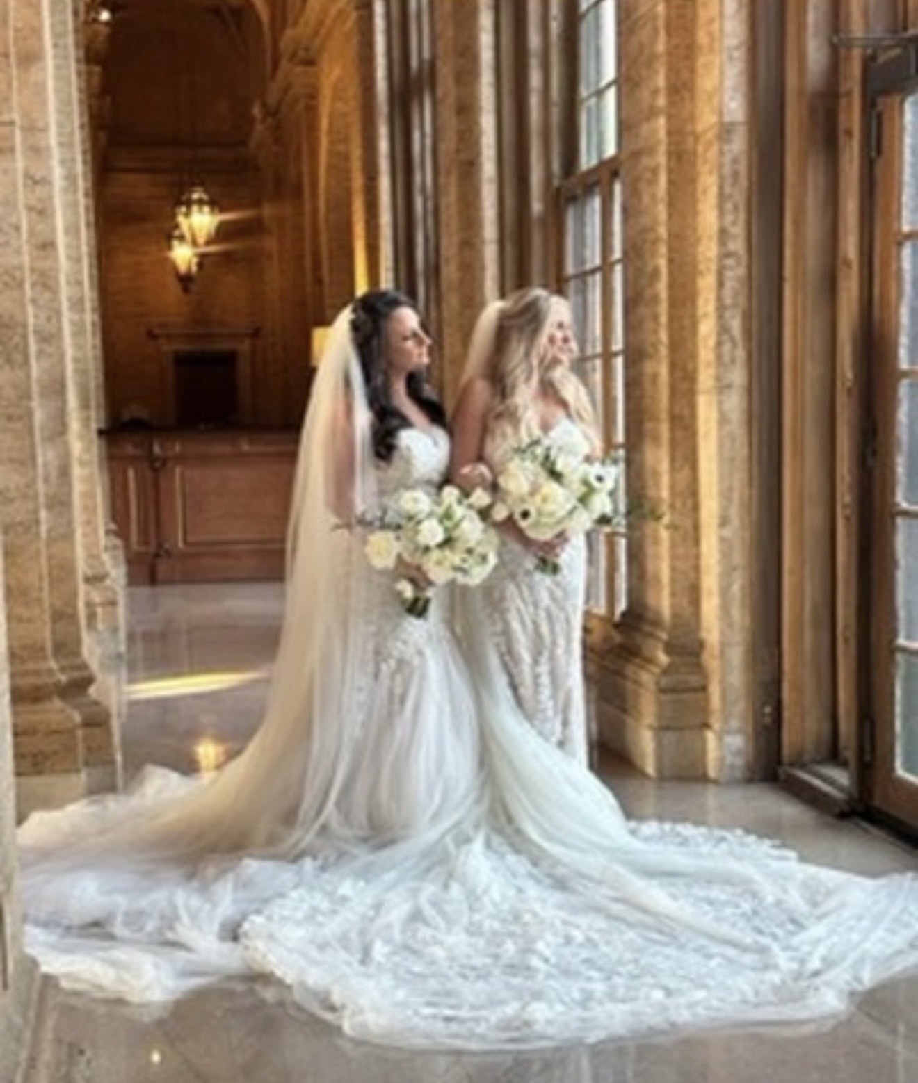 Two women in wedding dresses holding bouquets, standing by large windows in a grand, wood-paneled room.