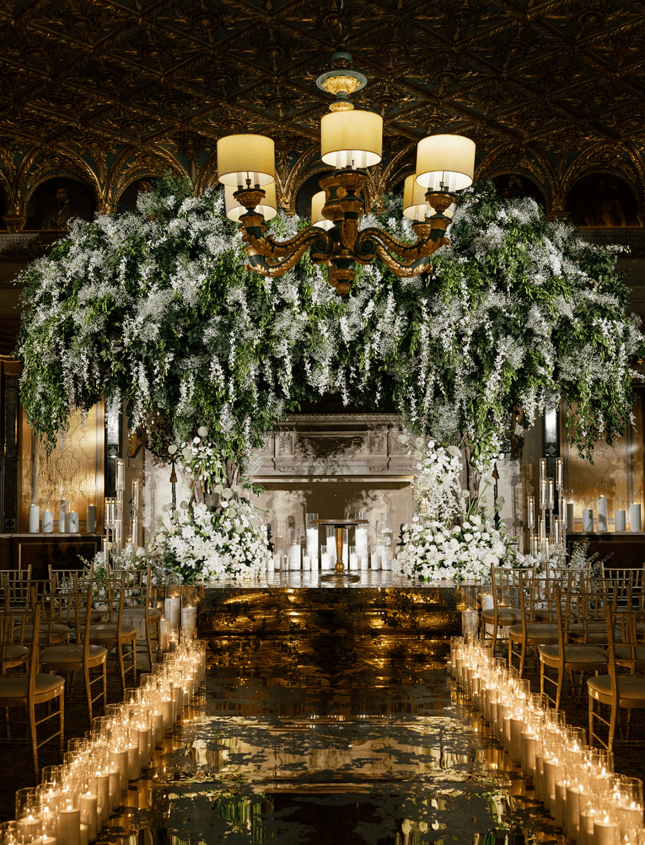 Elegant indoor setting with a chandelier, floral arrangements, candles, and a reflective aisle, likely for a wedding or event.