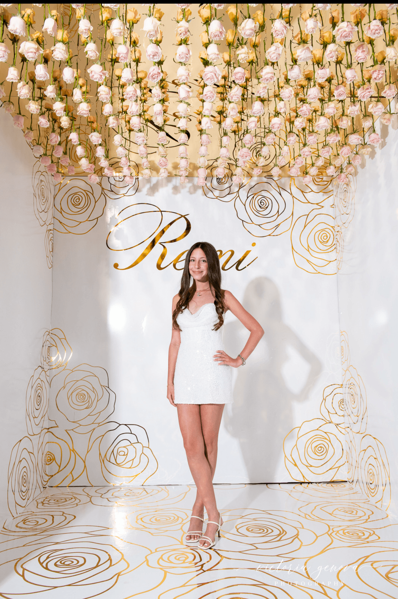 Woman in white dress standing on decorated stage with floral backdrop and hanging flowers, posing with hand on hip.