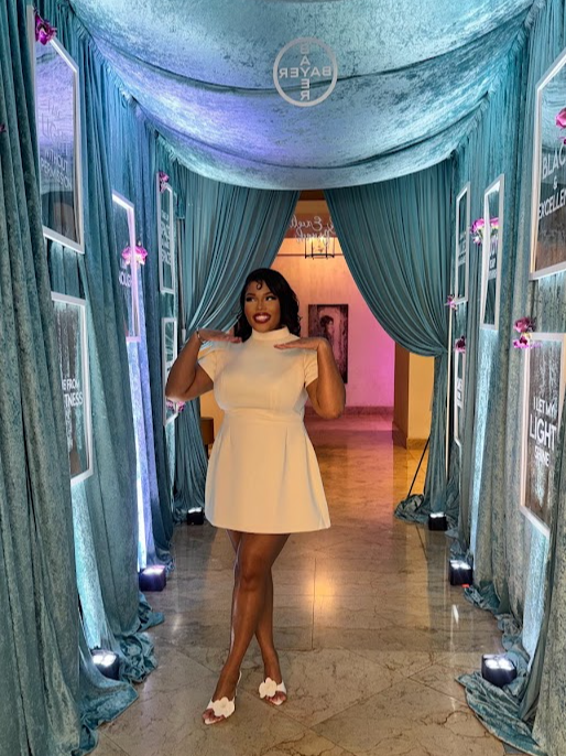 Woman in white dress standing in decorated doorway with blue curtains and pink lighting, smiling, holding her hair, wooden floor.