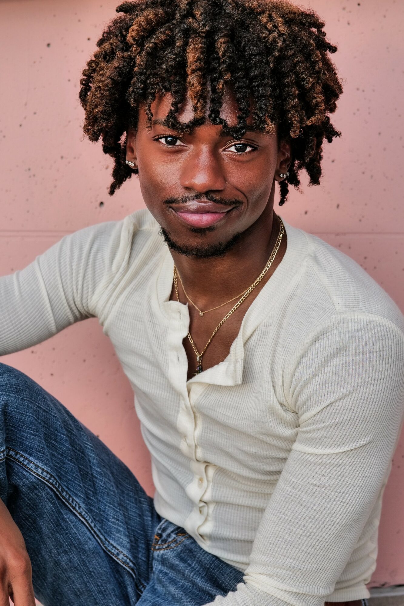 Young man with curly hair and a slight smile, wearing a white shirt and gold necklace, sitting against a pink background.