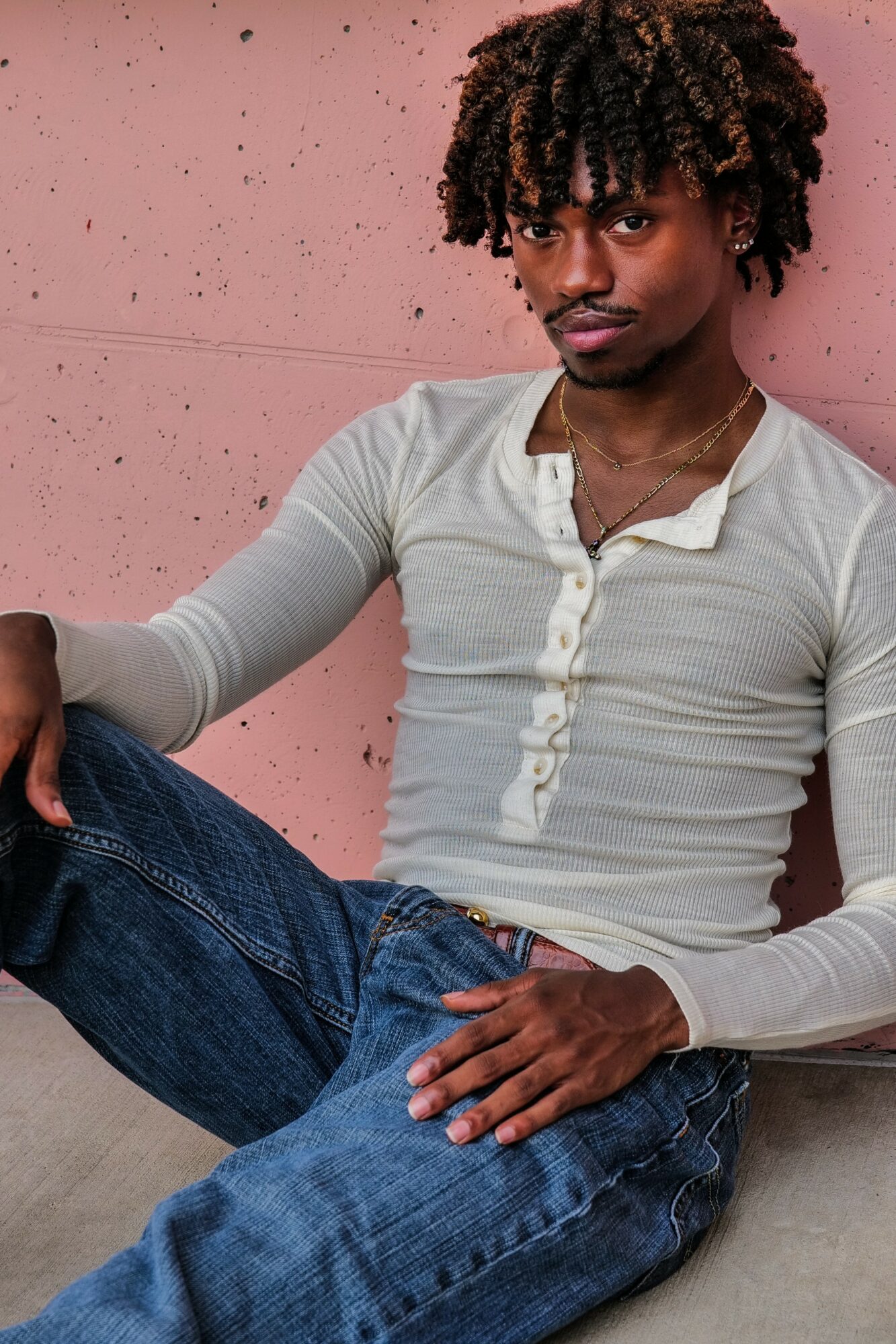 Young man with curly hair sitting against a pink wall, wearing a light-colored long-sleeve shirt and jeans.
