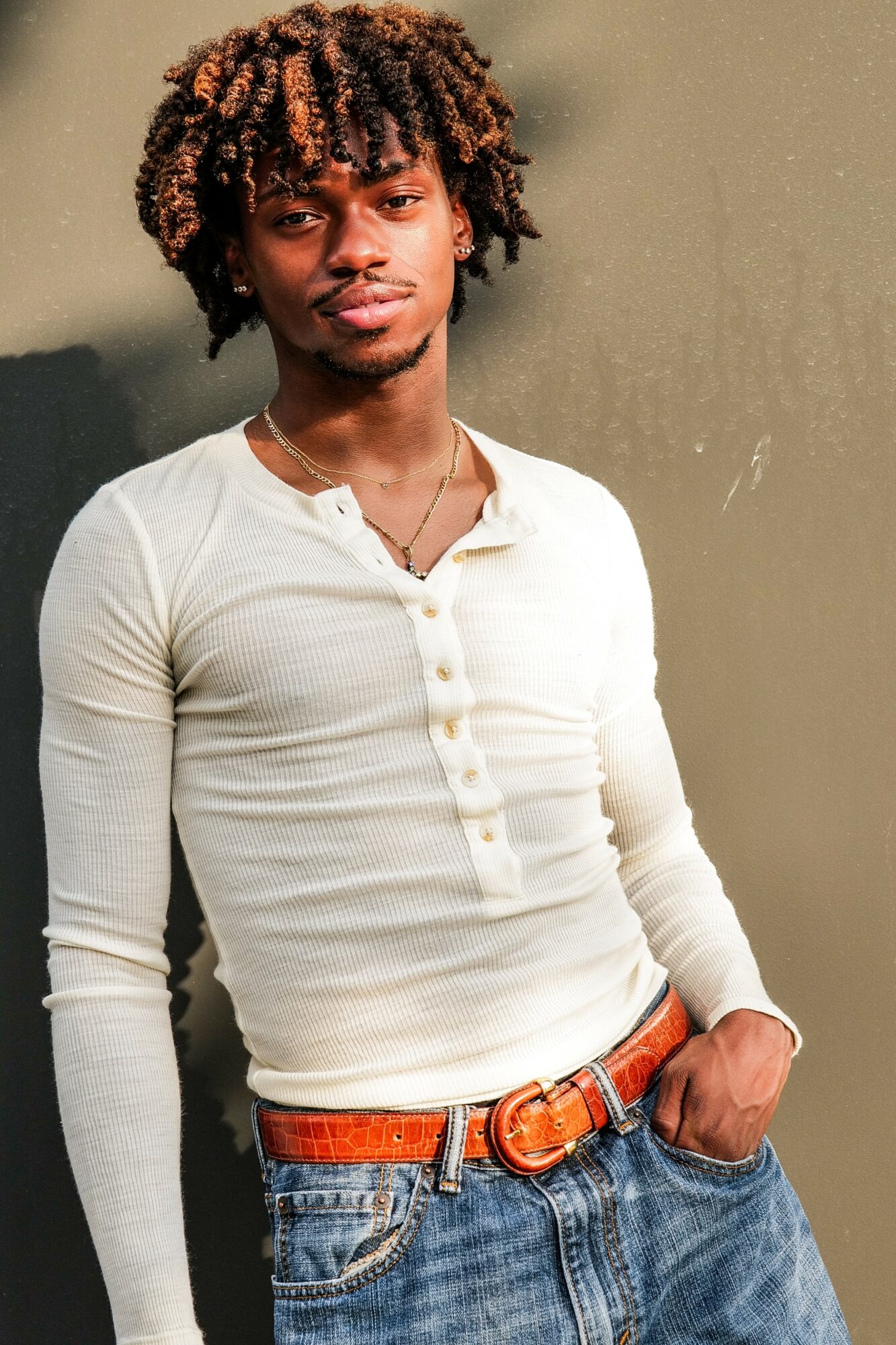 Young man with curly hair wearing a white long-sleeve shirt and jeans, standing against a plain wall.