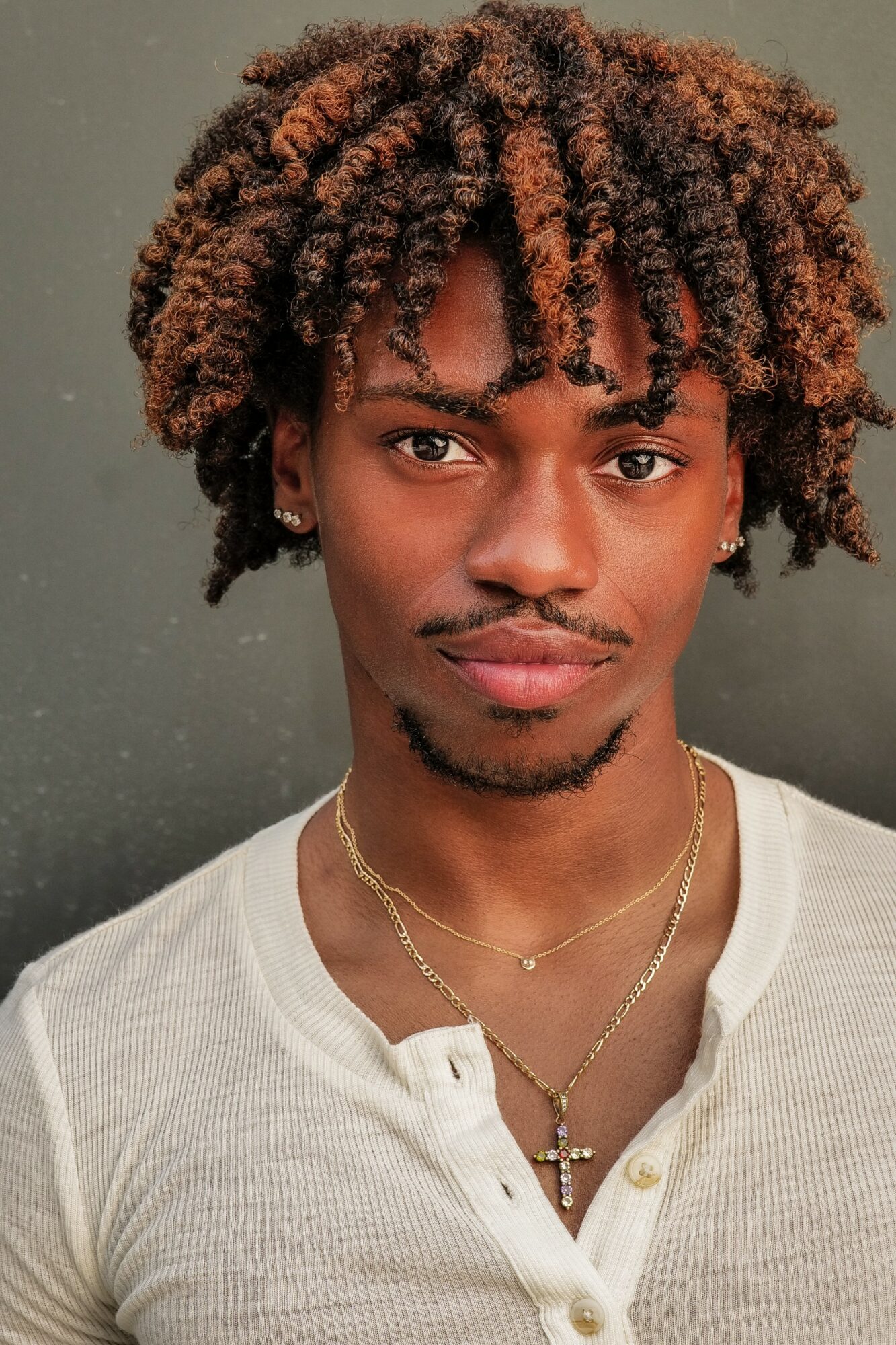 Young man with curly hair, wearing a white shirt and layered necklaces, looking at the camera.