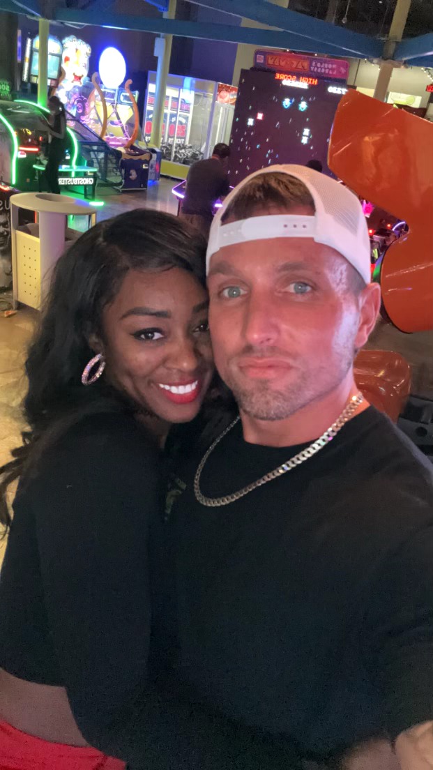 A man and woman taking a selfie at an amusement park, smiling with colorful rides in the background.
