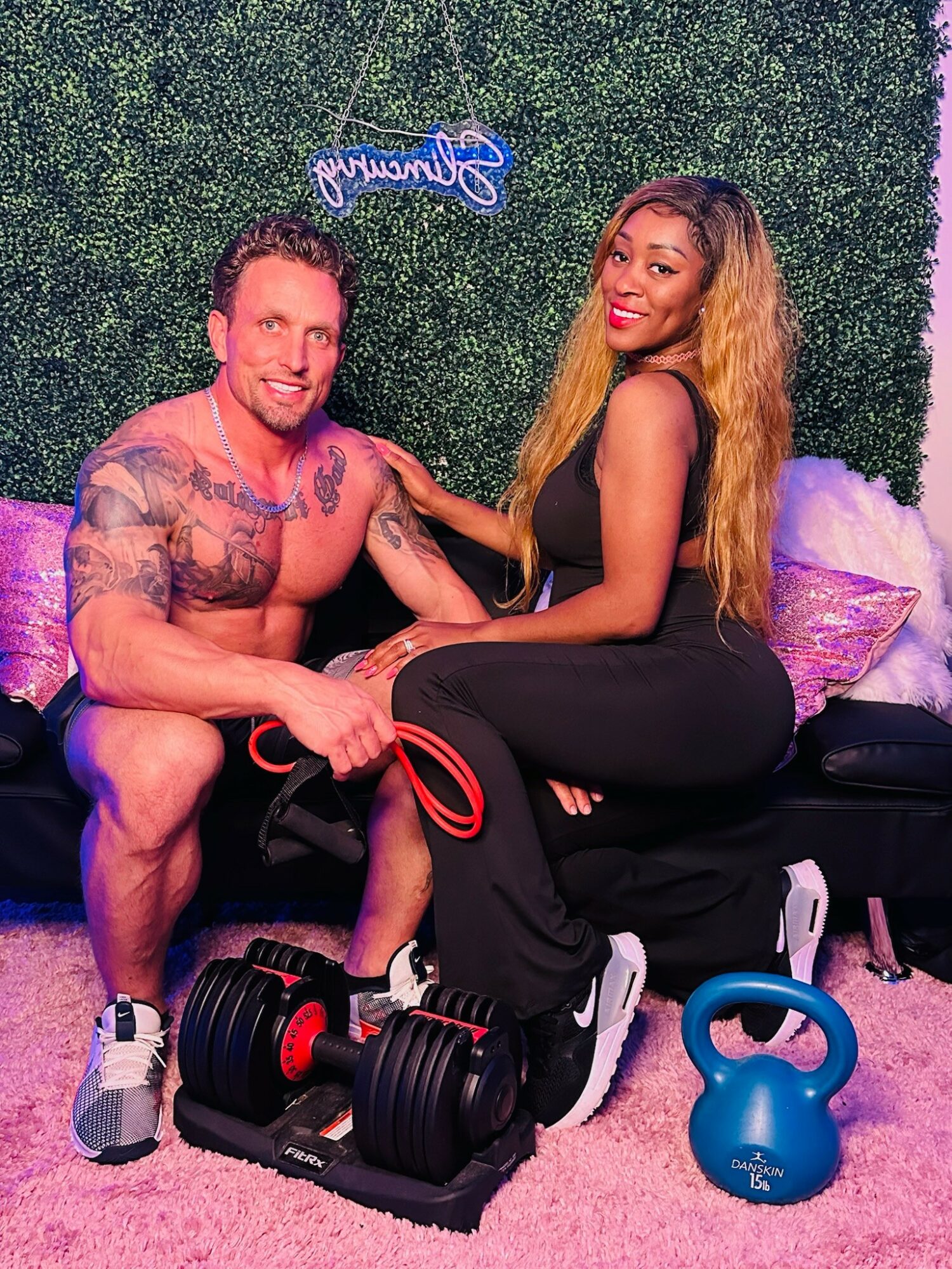 A man and woman sit together on a black bench, smiling, with fitness equipment and a green wall background.