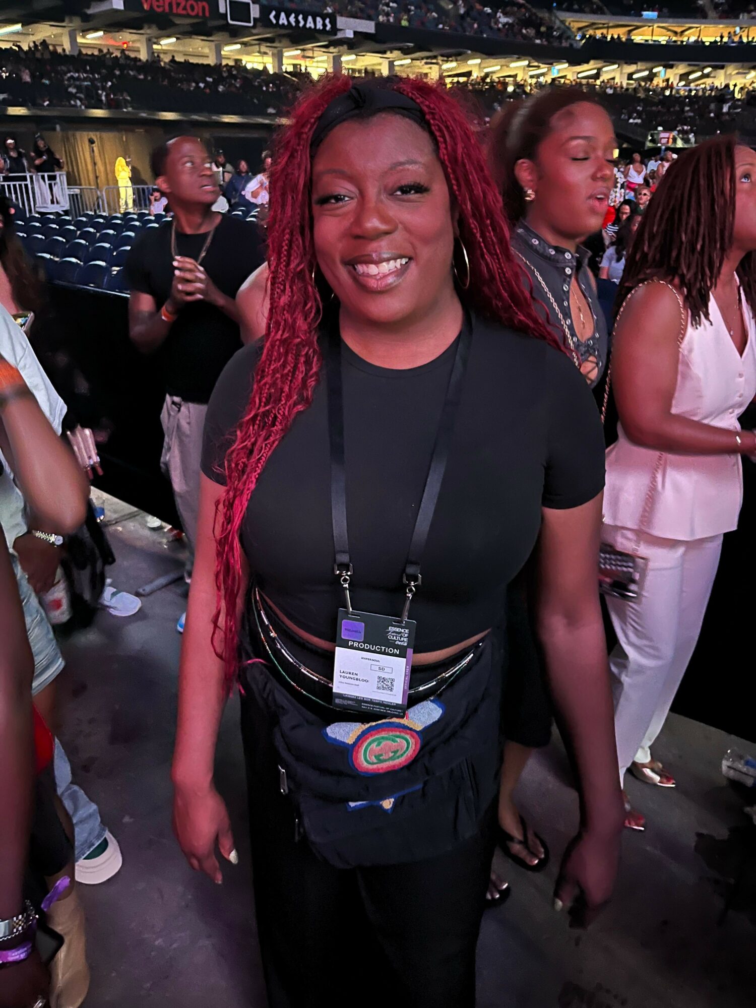 Smiling woman with long red braids wearing a black shirt and conference badge at an event, with people and city lights in background.
