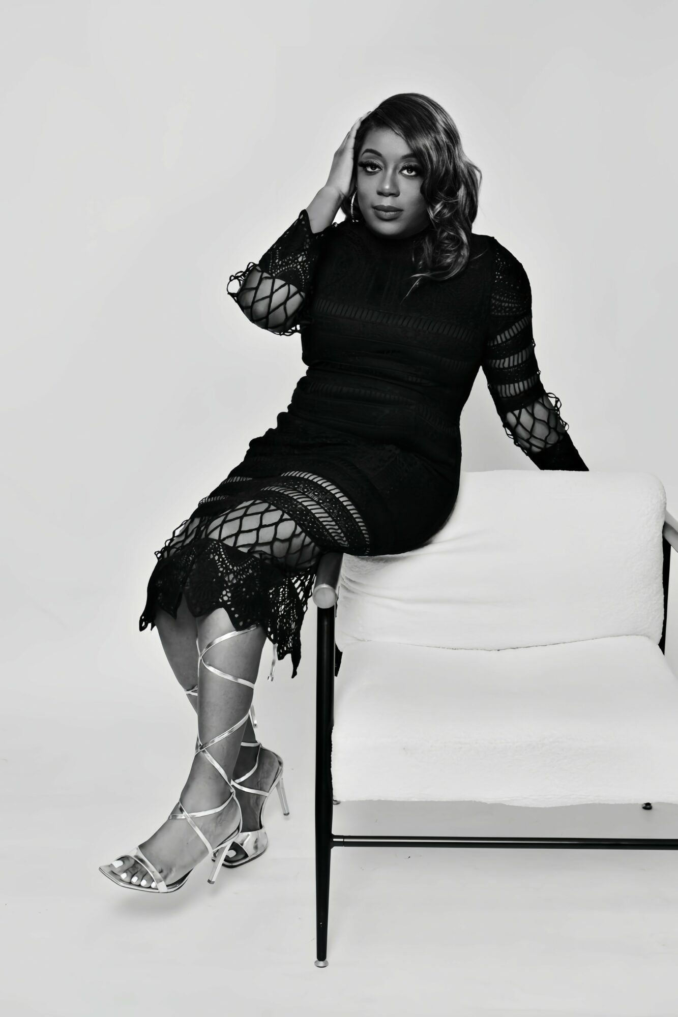 Woman sitting on a chair, wearing a black dress with lace details, posing with her hand on her head, in black and white.