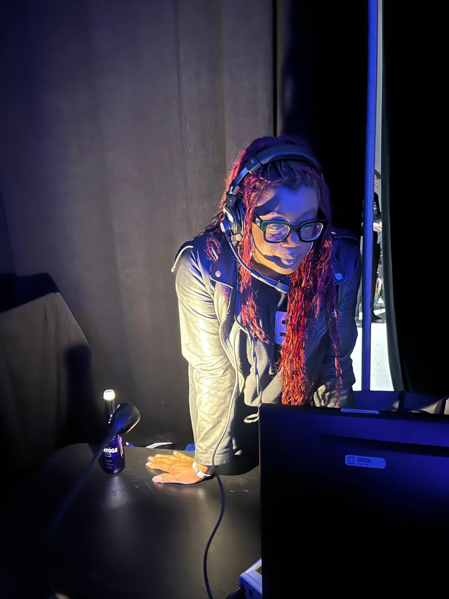 Person wearing headphones and glasses working on a laptop in a dark room, with a water bottle nearby.