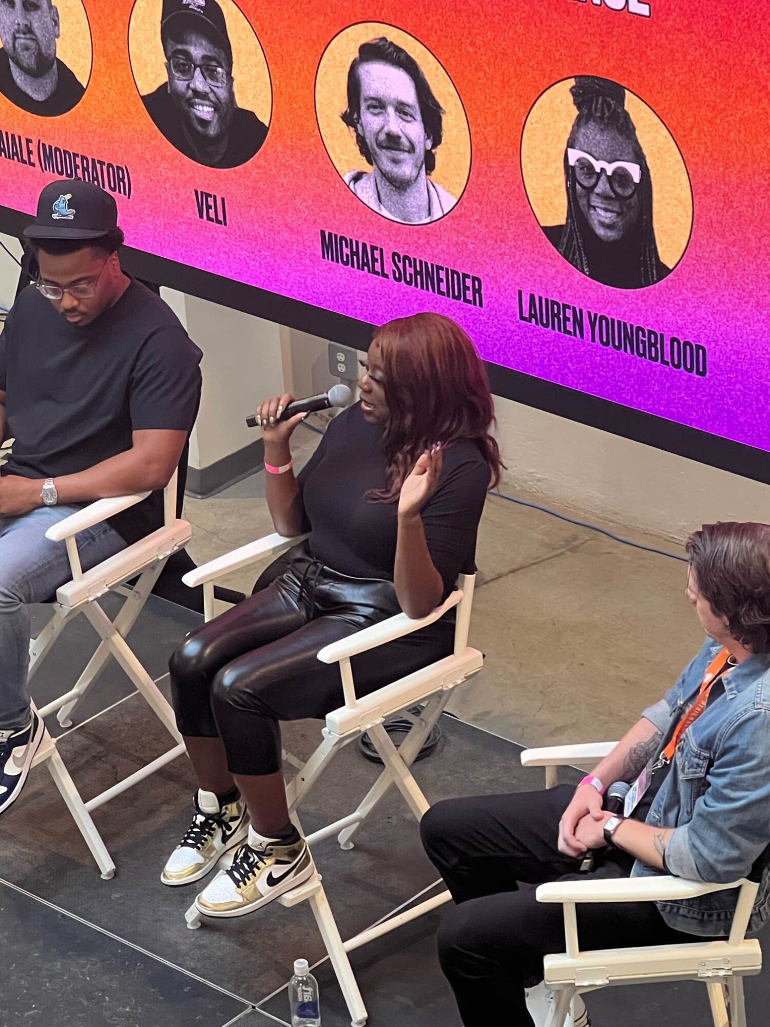 Woman with red hair speaking into a microphone, seated on a white chair, with two people nearby, in front of a colorful screen.