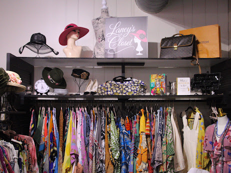 Clothing and accessories displayed on shelves and racks in a boutique, with hats, bags, and colorful dresses.