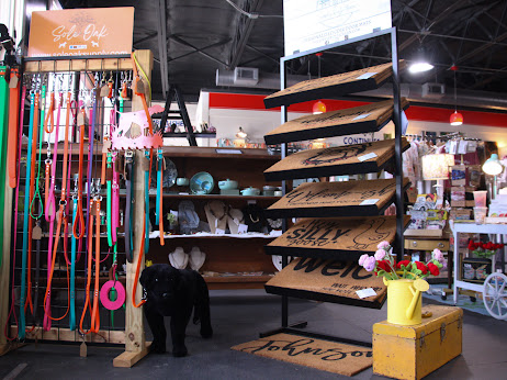 Display of colorful pet leashes and collars, black dog near shelves, store interior with various items, yellow watering can with flowers.