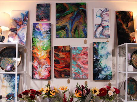 Gallery wall with colorful abstract paintings, two white shelves with decorative items, and a lamp on the left. 20 words.
