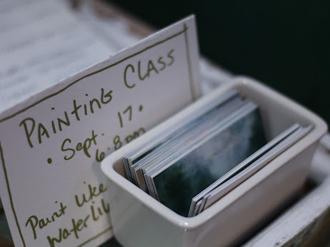A white container holding small cards, with a handwritten sign reading 'Painting Class' and date, on a table.