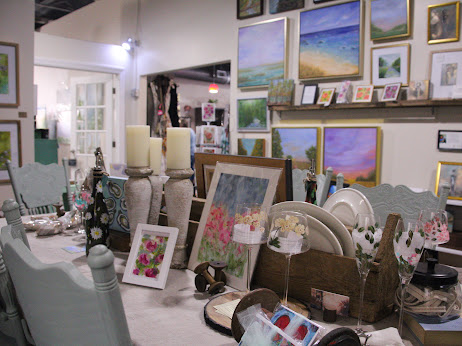 Interior of an art gallery with paintings, framed photos, and decorative items on tables and shelves.