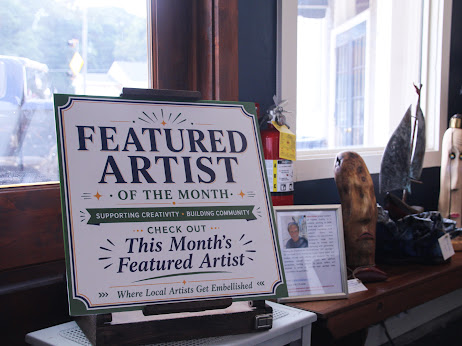 Signboard displaying featured artist of the month, supporting community, with a check-out message, placed on a windowsill with decorative items.
