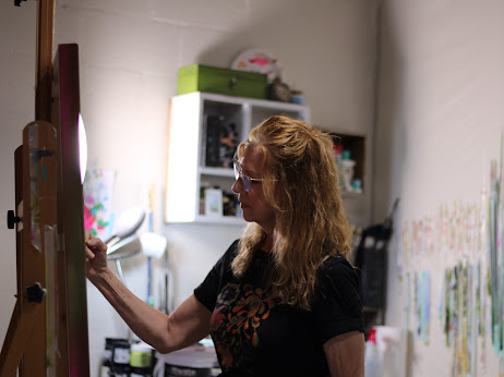 Woman with curly hair painting on an easel in a room with shelves and colorful artwork