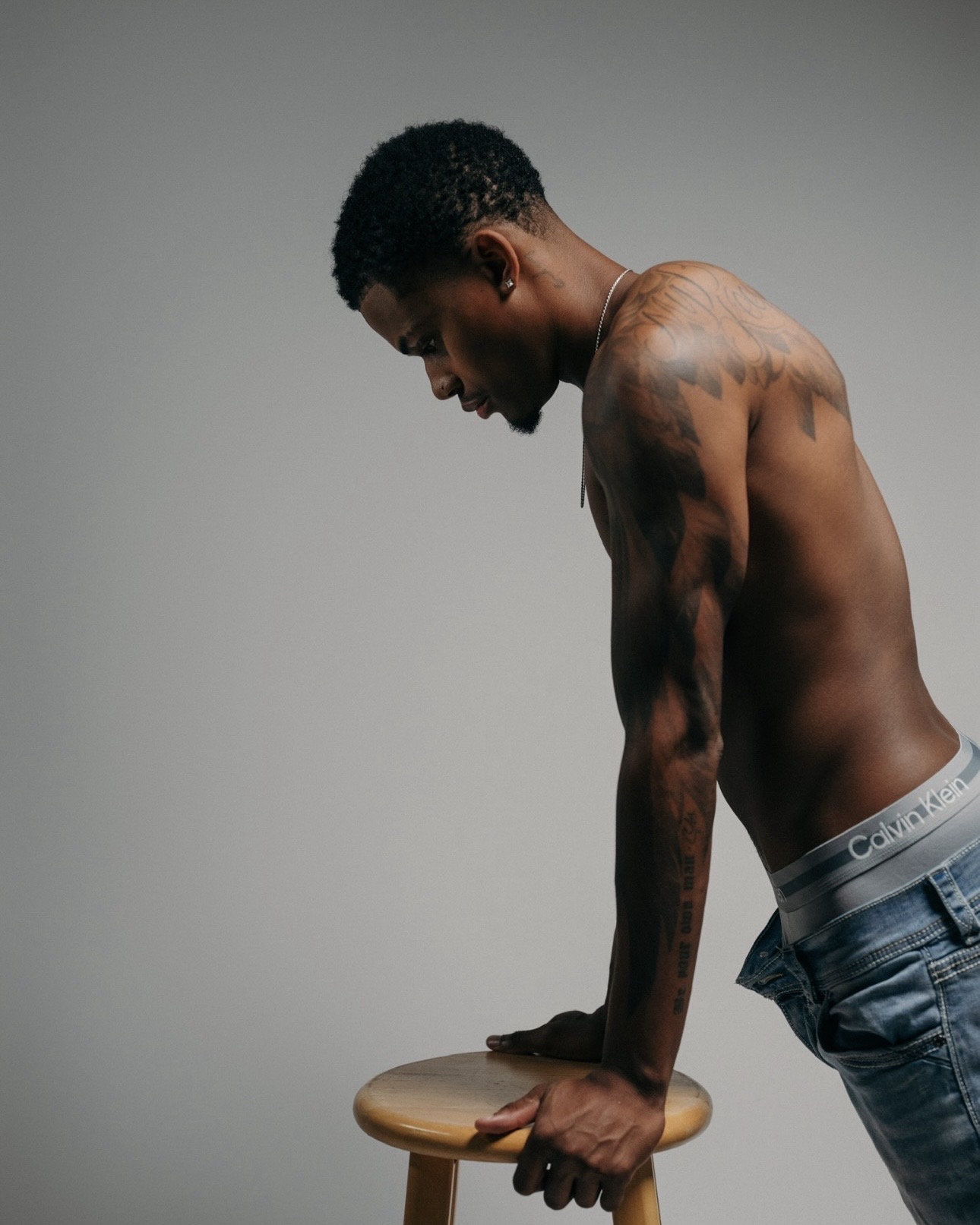 A shirtless man with tattoos leans forward on a wooden stool, wearing jeans, against a plain gray background.