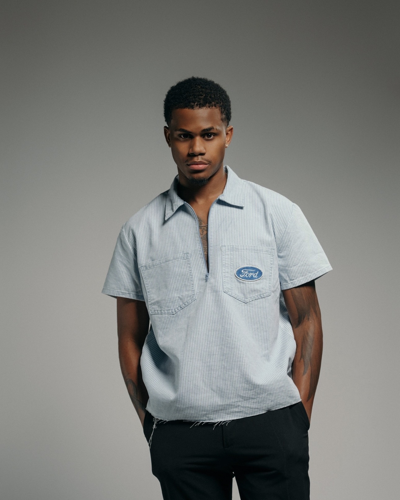 Young man with short curly hair wearing a light gray polo shirt and black pants, standing against a plain background.