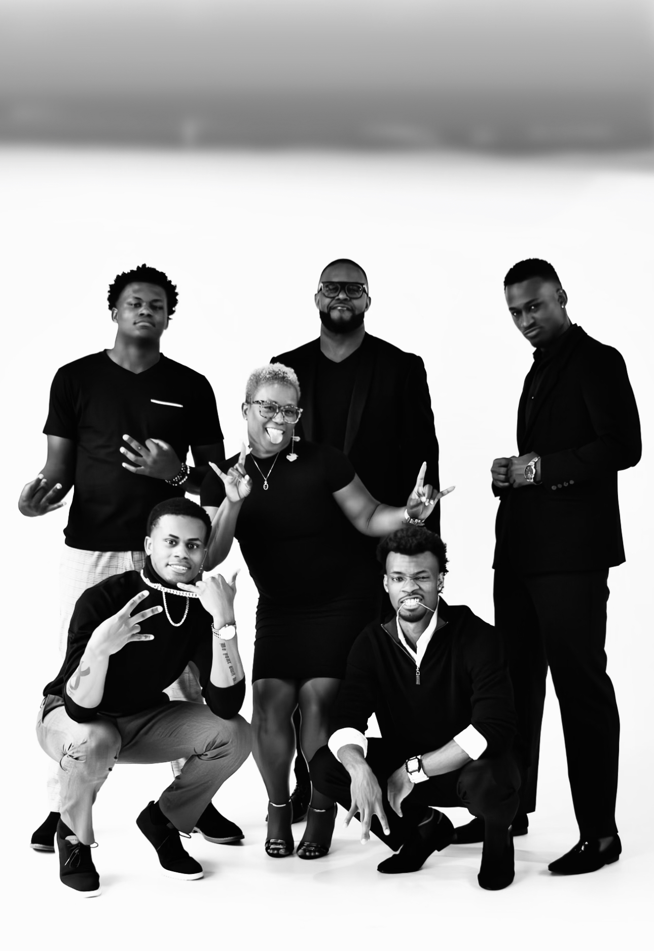 Group of six people posing together, some standing and some crouching, in black clothing, smiling and making gestures, against a plain background.