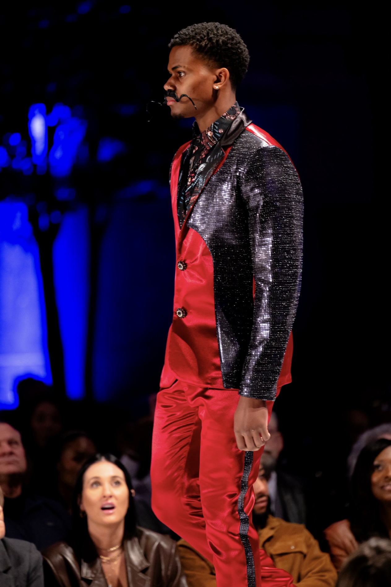 Man walking on runway wearing red and black outfit, audience watching, dark background with blue lighting, side view