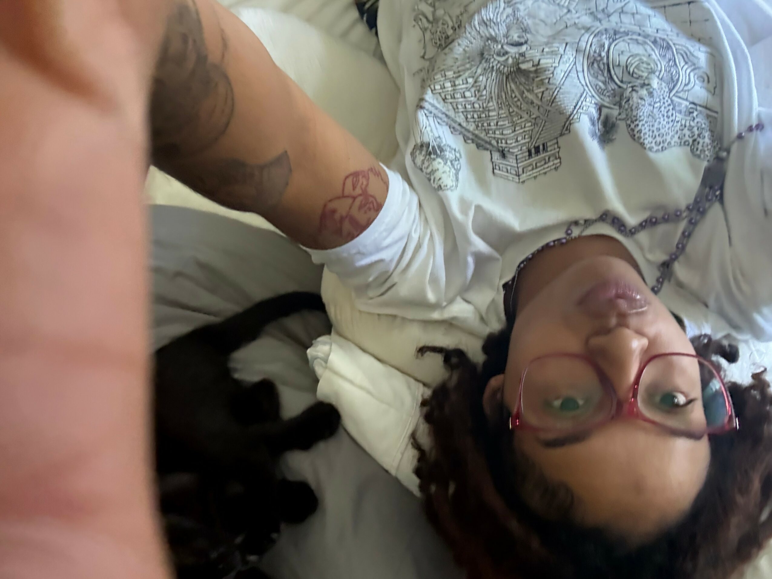 Person lying on bed with glasses, tattoos, and braided hair, holding a black and white dog, in a relaxed setting.