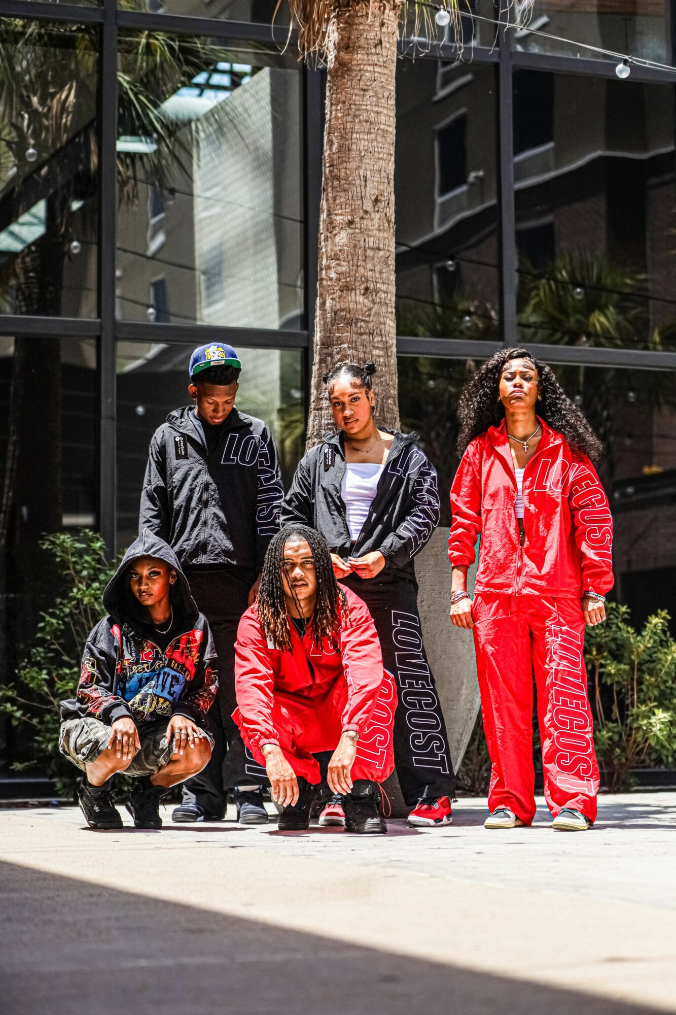 Five people pose outdoors in front of a modern glass building, some wearing red and black clothing, with trees and plants nearby.
