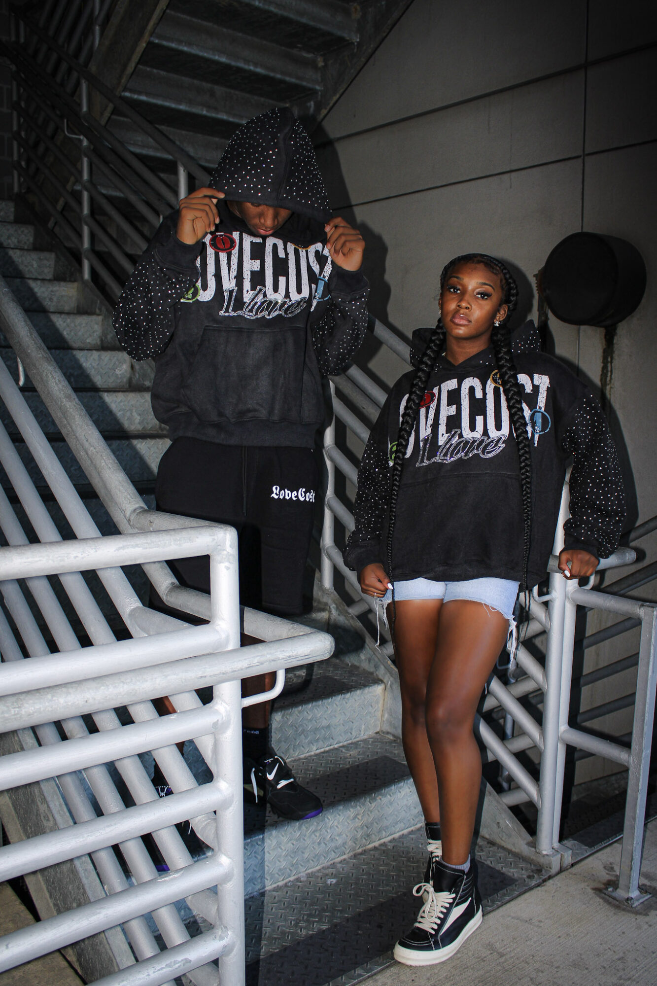 Two young women in black hoodies with white text, one with a hood over her head, standing on stairs indoors.