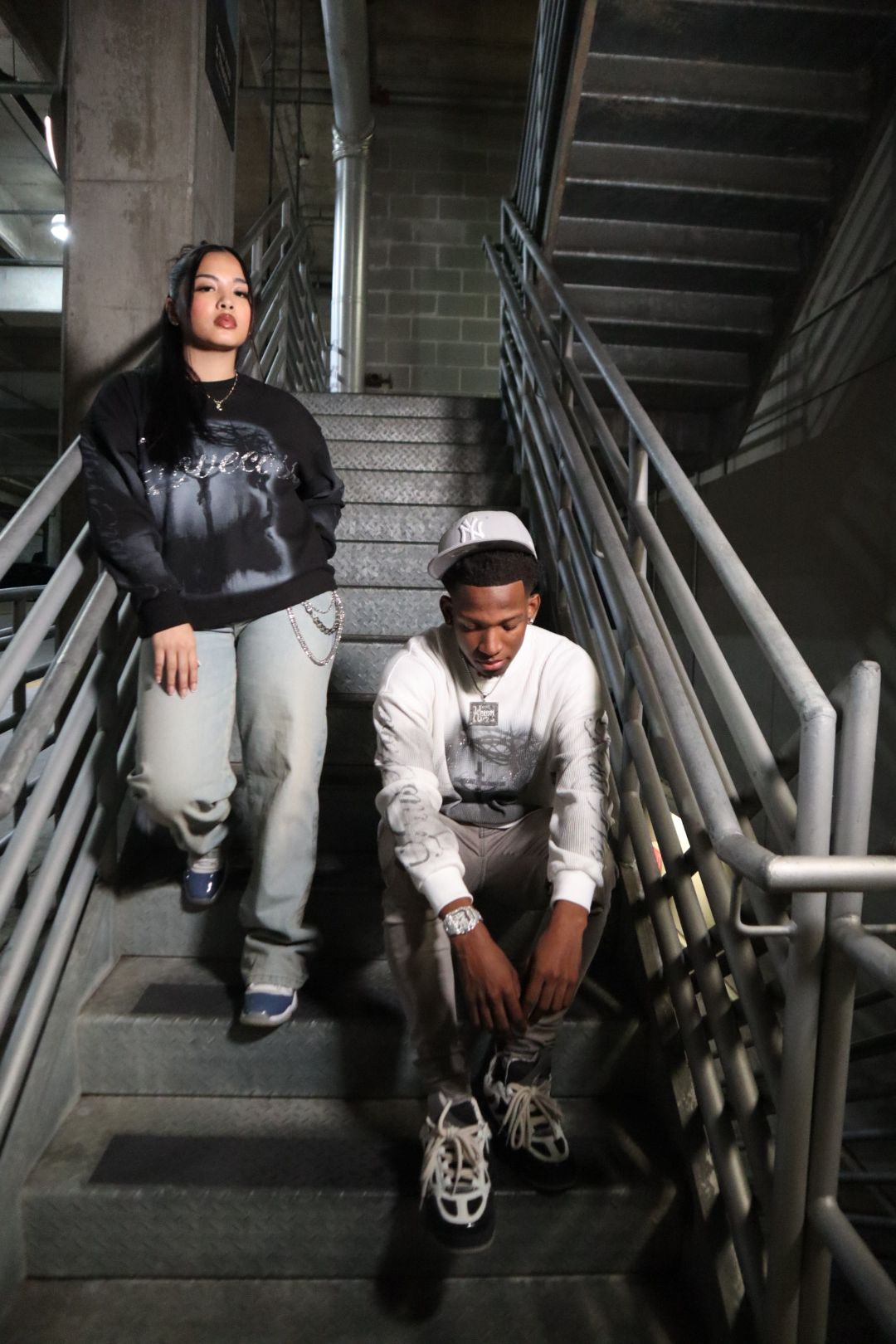 Two people walking down a staircase in an industrial setting, one standing and one sitting on the stairs, with metal railings.