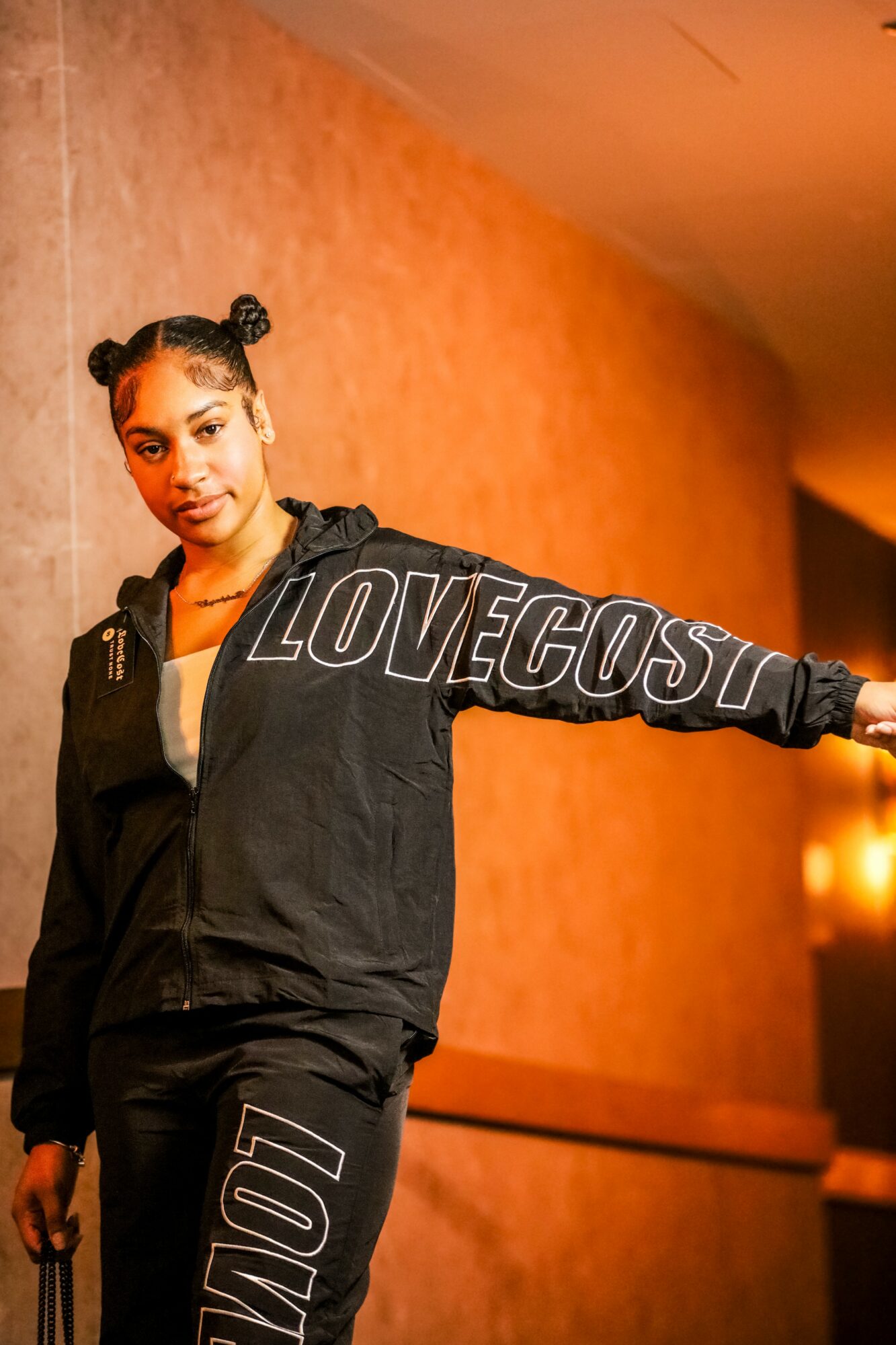 Young woman with two buns hairstyle wearing a black outfit and a black jacket with 'LOVECOST' written on the sleeve, standing indoors against a warm-colored wall.