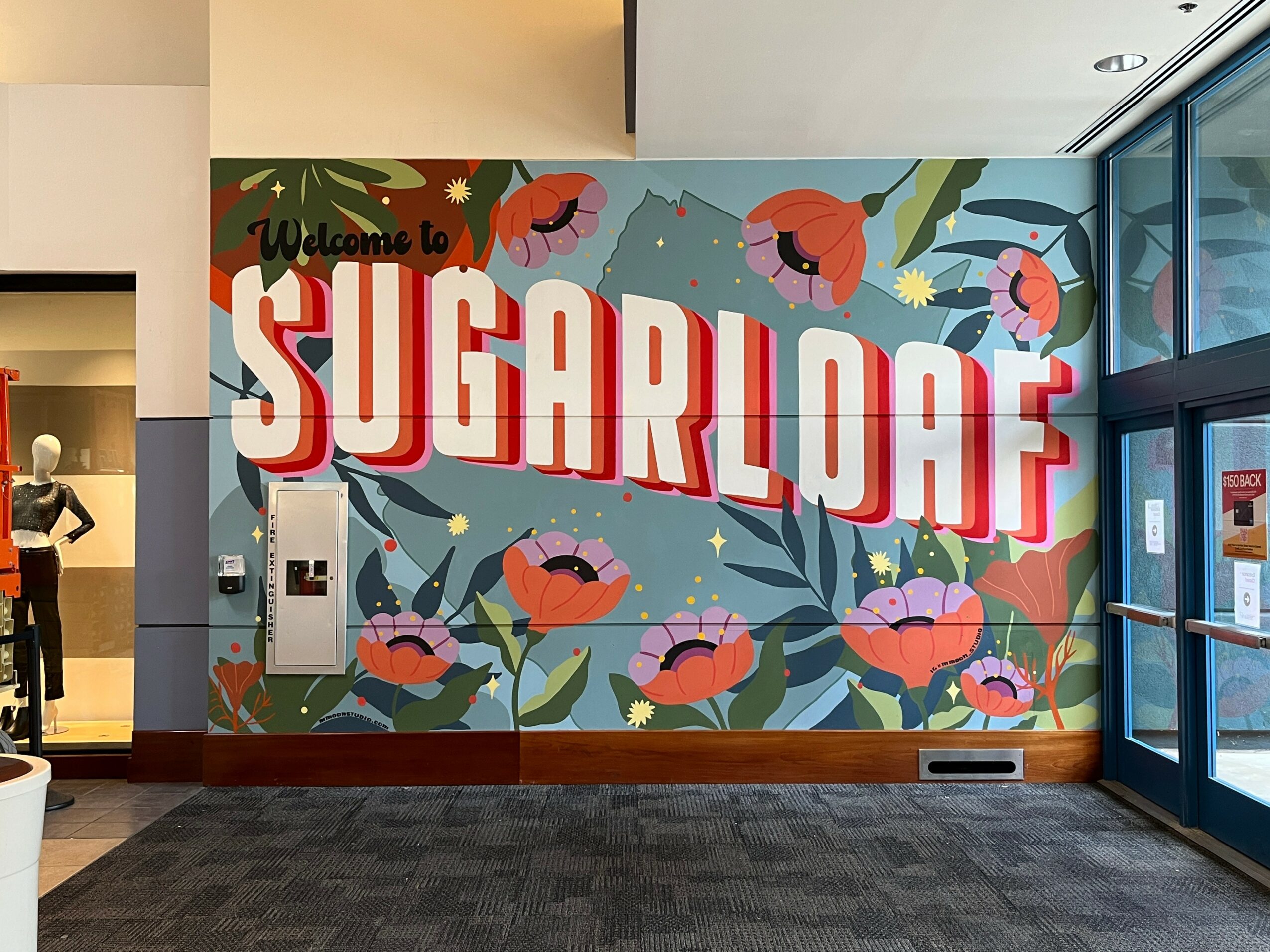 Colorful wall mural with large text 'SUGARLOAF' and floral design, welcoming visitors, with a small sign and a person in the background.