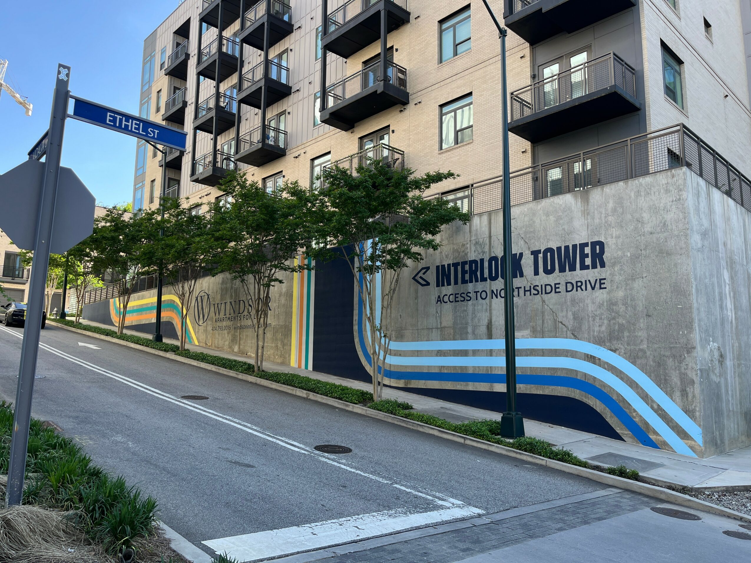 Street view of a building with a mural, trees, and a sign for Interlok Tower, with a crosswalk and sidewalk.