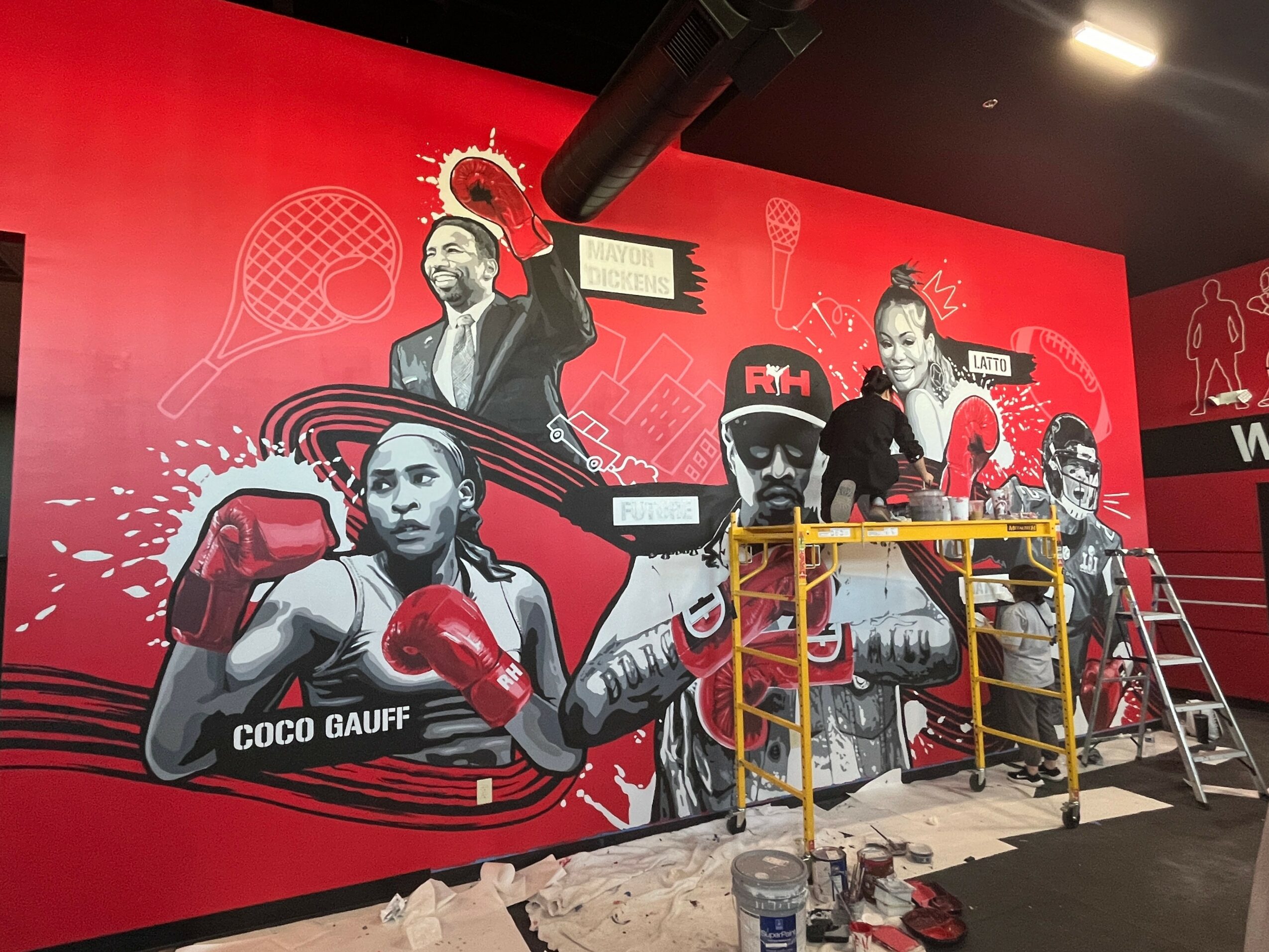 Colorful mural of boxers and athletes with sports equipment on red background, with scaffolding and tools in front.