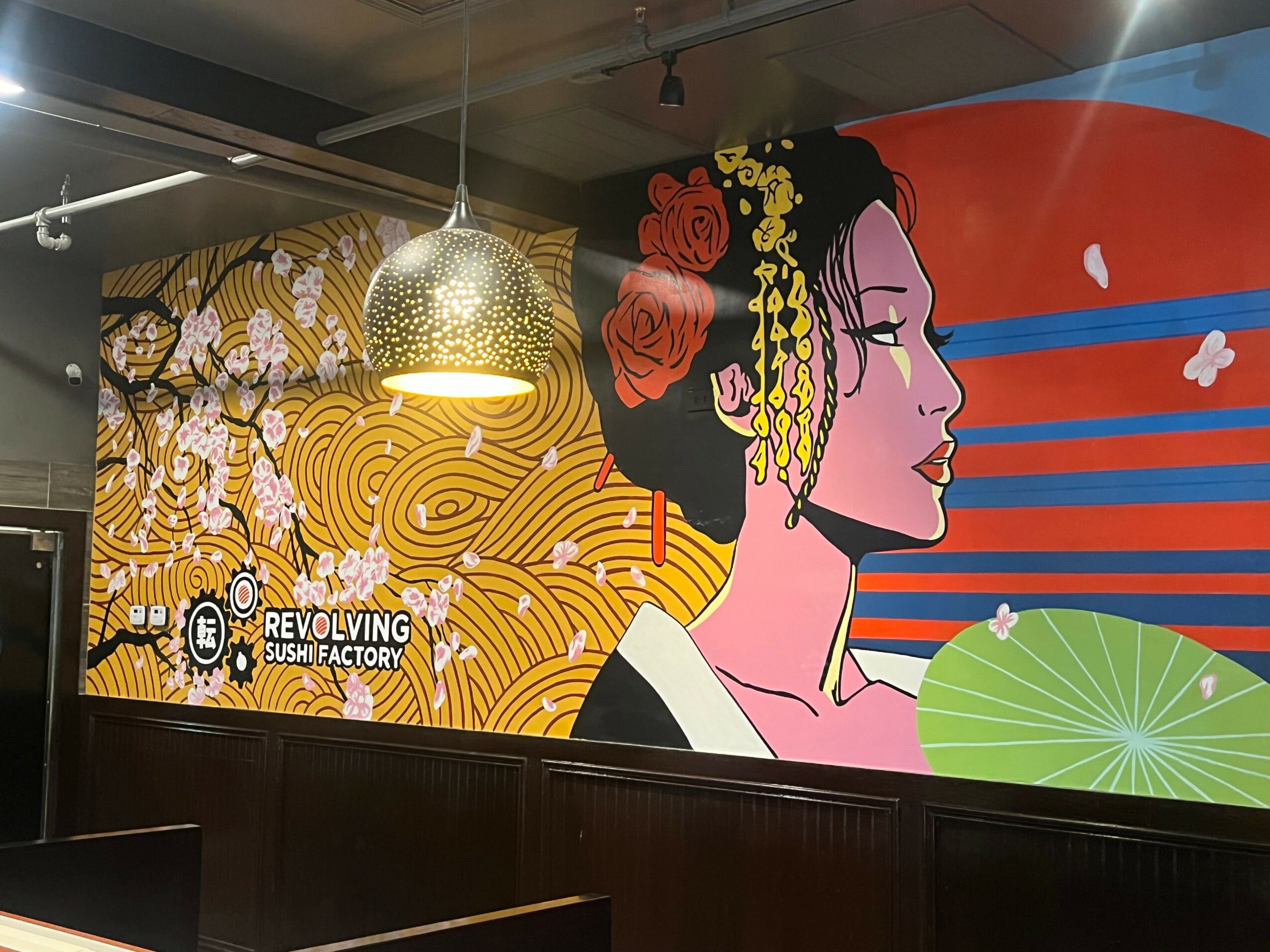 Colorful mural of a woman with traditional Japanese hairstyle and makeup, with a red sun, blue stripes, and a green fan.