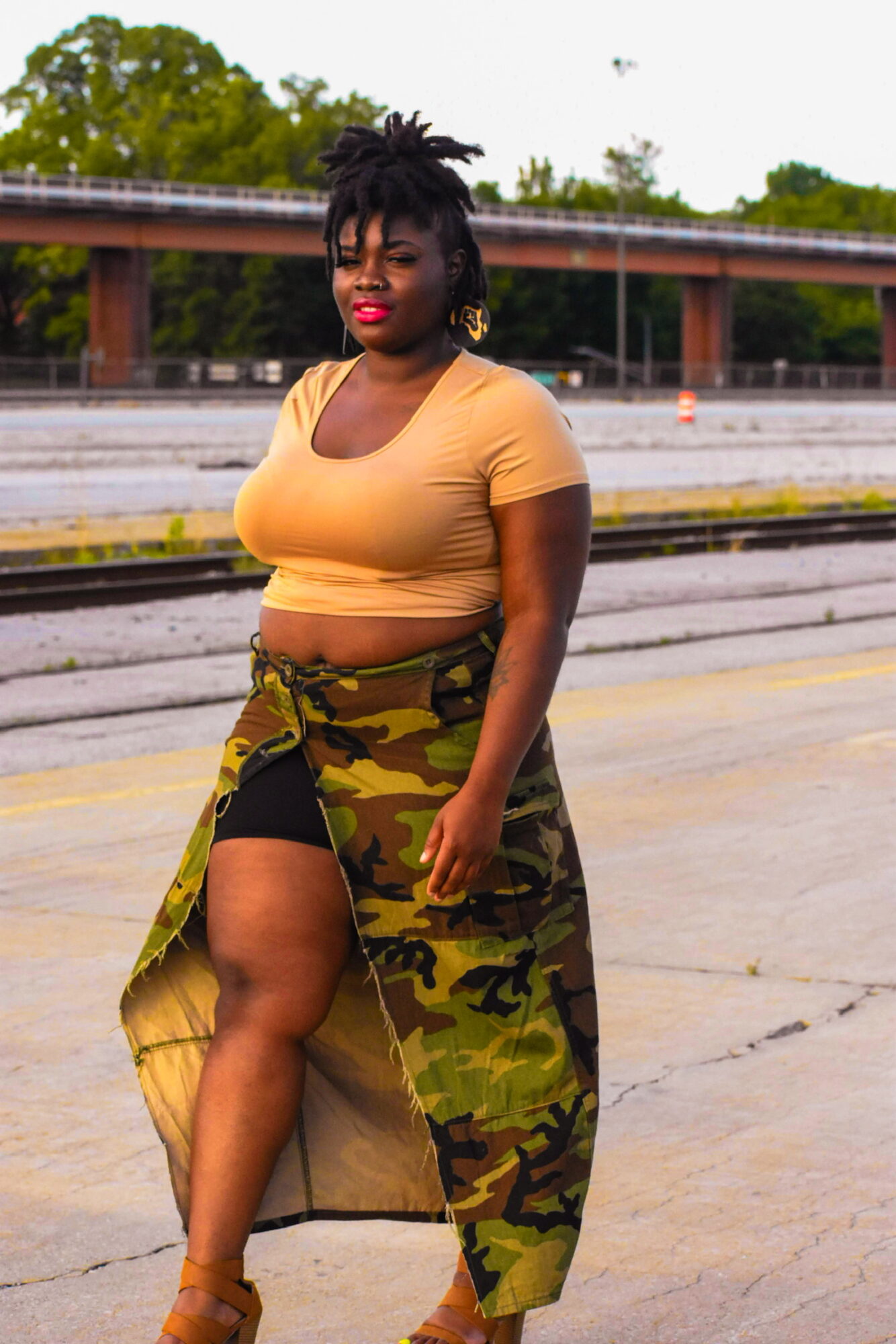 Woman with dark hair and red lipstick standing outdoors on a street, wearing a yellow top and camouflage skirt, with a bridge in background.