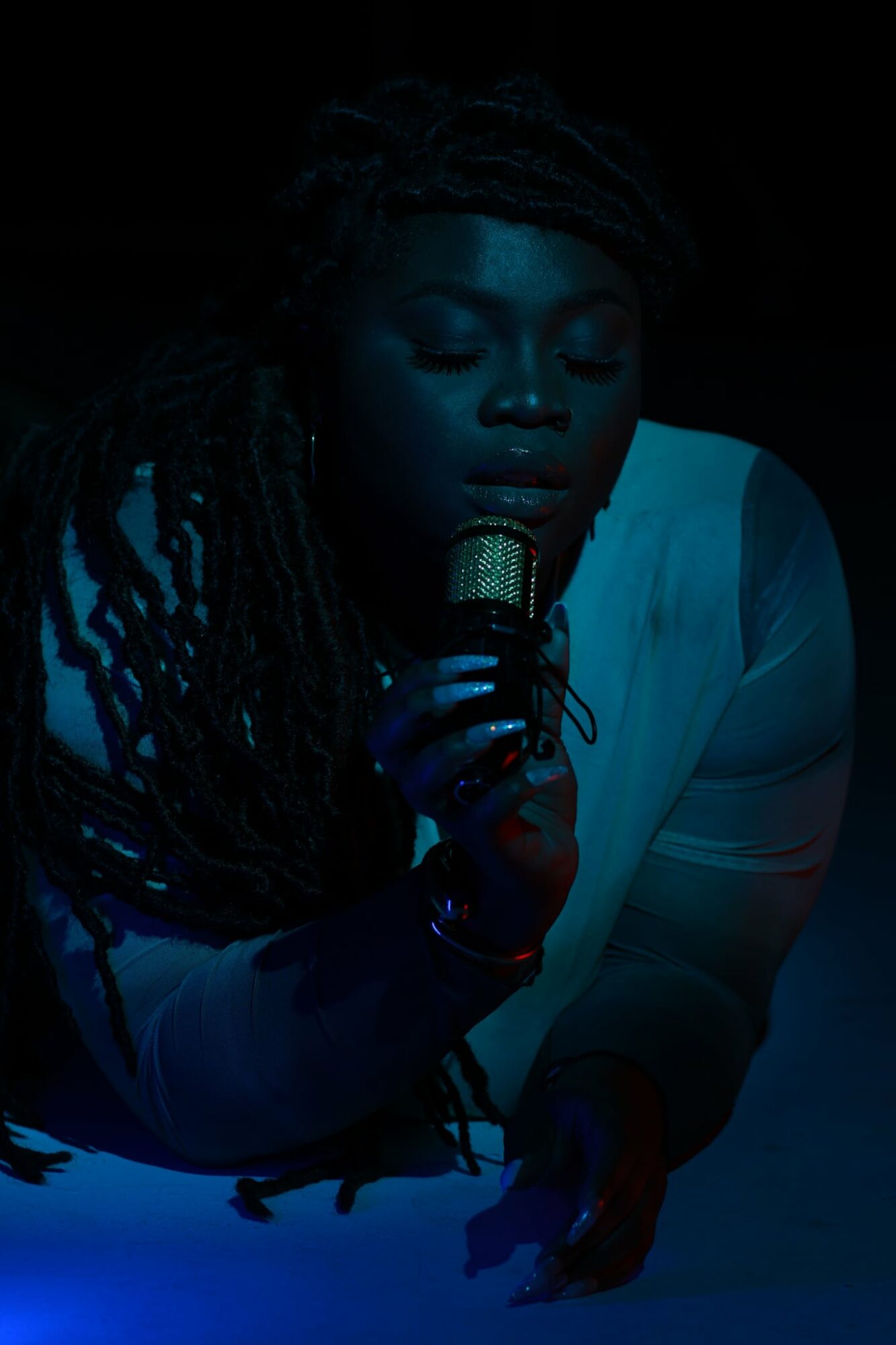 Person with dreadlocks holding a microphone, eyes closed, in a dark setting with blue lighting.