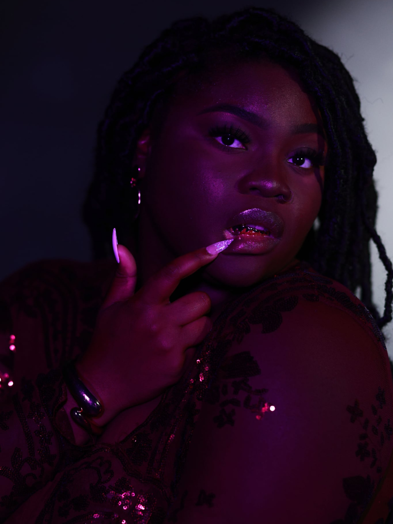 Portrait of a woman with dark skin and long dreadlocks, touching her lips, in low lighting with purple tones.