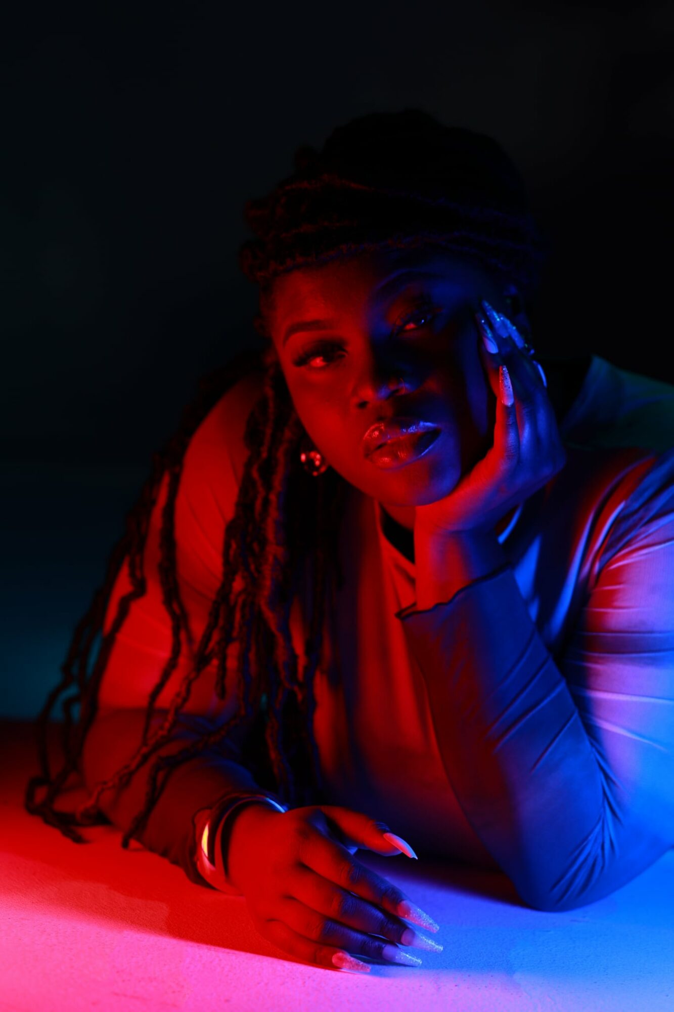 Person with long dreadlocks resting chin on hand, illuminated by red and blue lighting, looking at camera.